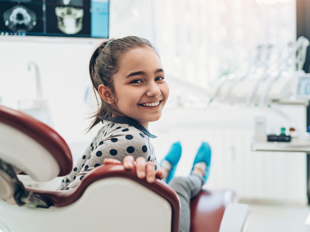 Little girl looking behind her at the camera and smiling while sitting in a dental chair.